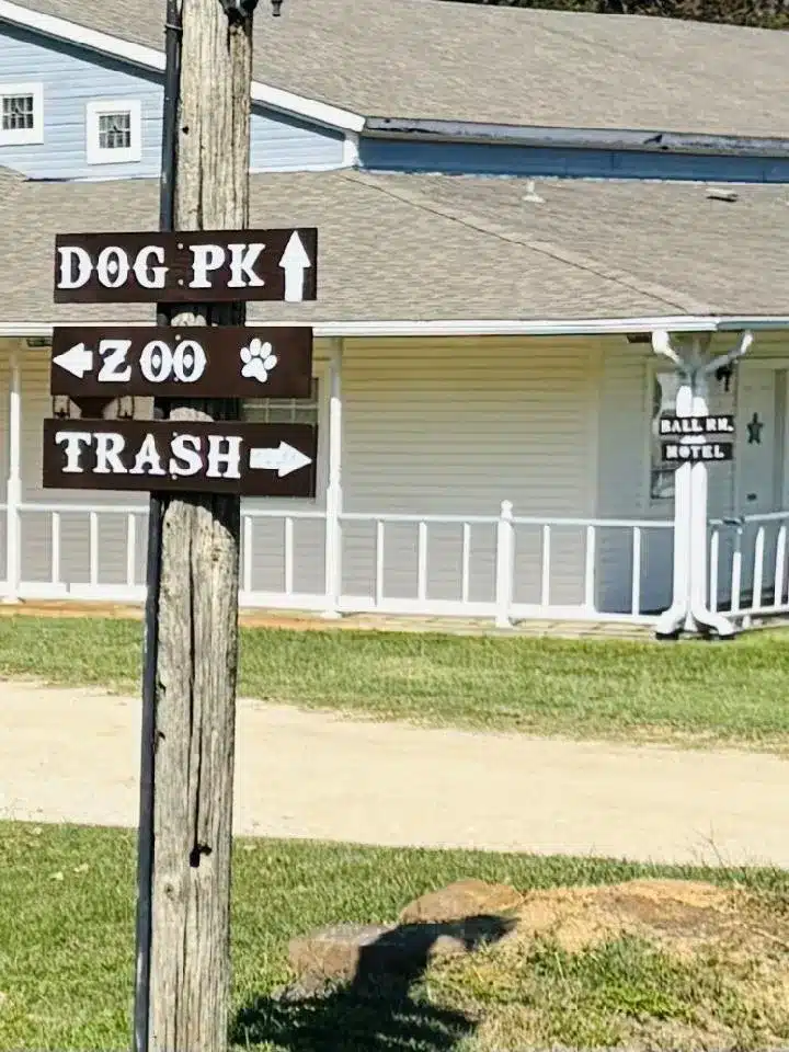Wooden utility pole with rustic directional signs reading Dog Pk, Zoo, and Trash in front of a white-porched building.