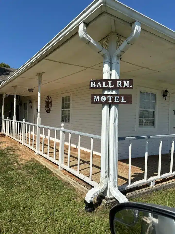White single-story motel building with covered wooden porch, railing, and a brown "Ball Rm. Motel" sign on a post.