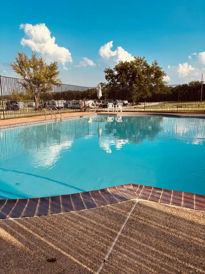 Large outdoor swimming pool with blue water, ladder handrails, fenced deck with white patio chairs, umbrella, and trees