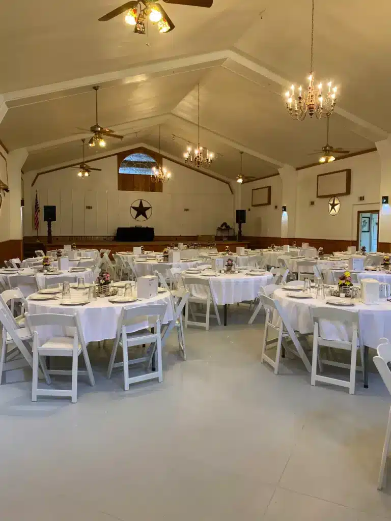 Interior of an event hall with round tables in white linens, folding chairs, chandeliers, ceiling fans, and a Texas