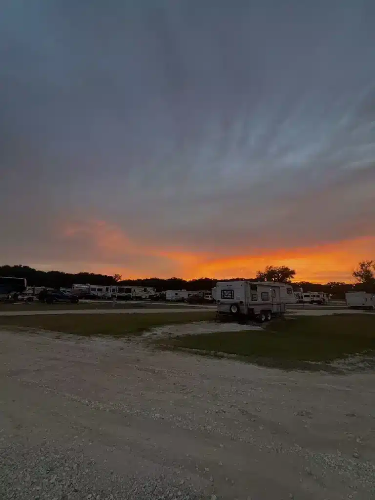 Fiery orange sunset behind tree line over an RV park with parked travel trailers, grass lawn, and gravel road in