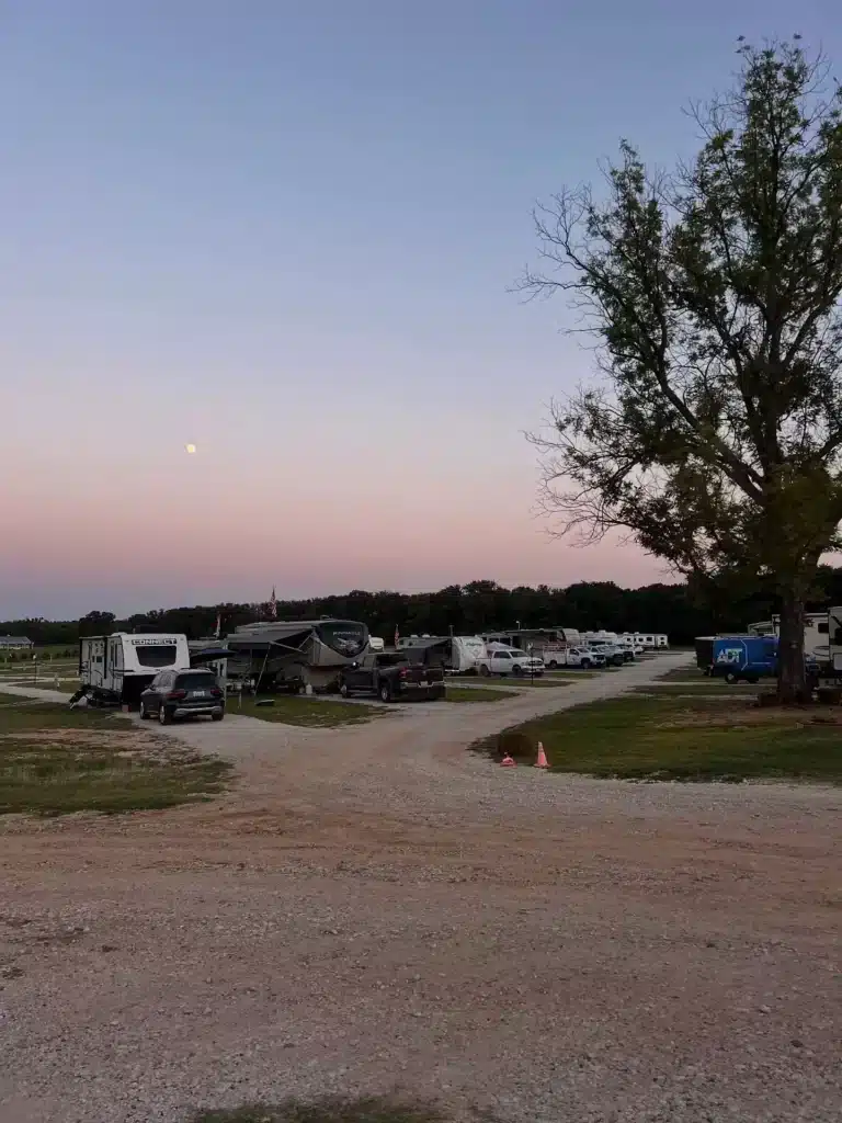 Gravel road through an RV park at dusk with travel trailers, trucks, an orange cone, and moon rising over pink sky.