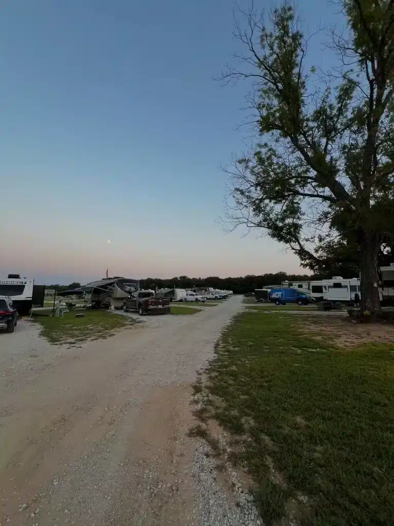 Gravel road through an RV park at dusk with parked fifth-wheel trailers, pickup trucks, a blue van, and large shade