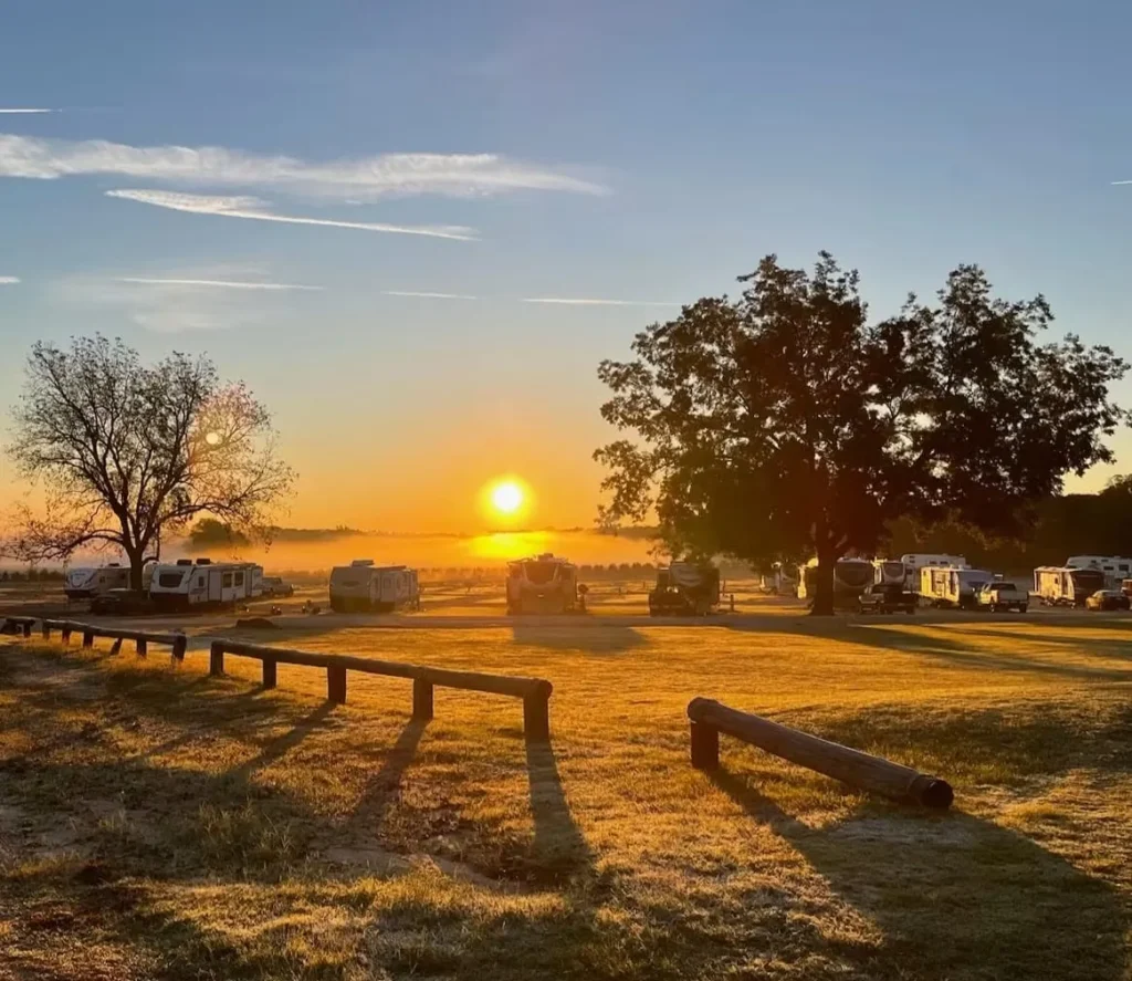 Golden sunrise over a grassy RV park field with parked travel trailers, large oak trees, and wooden log fencing casting