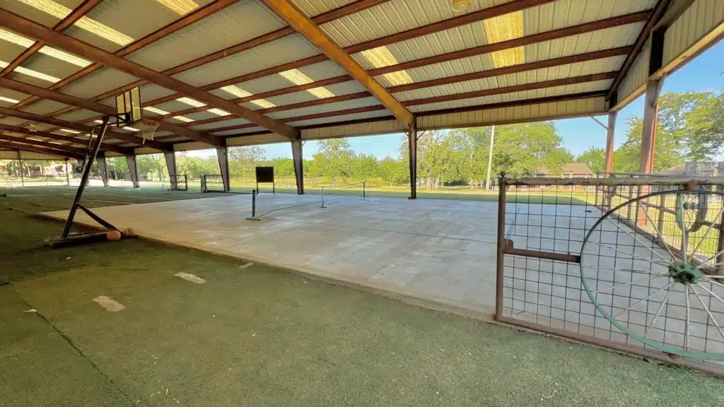 Large open-sided metal pavilion with concrete sport court, basketball hoop, pickleball nets, artificial turf edges, and