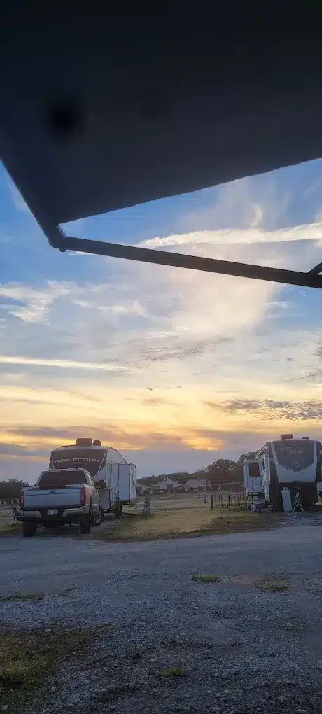 View from under an RV awning at sunset showing a white pickup truck, travel trailers, and gravel sites.